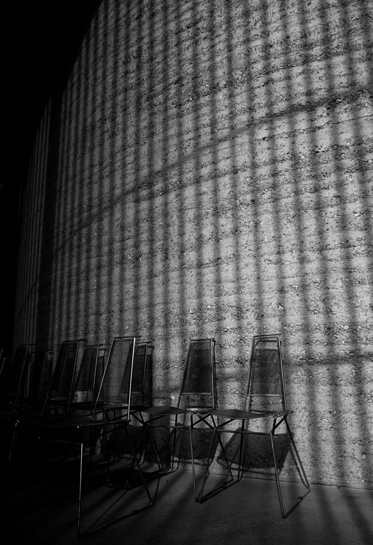 A row of metal chairs against a concrete wall striped with light and shadow