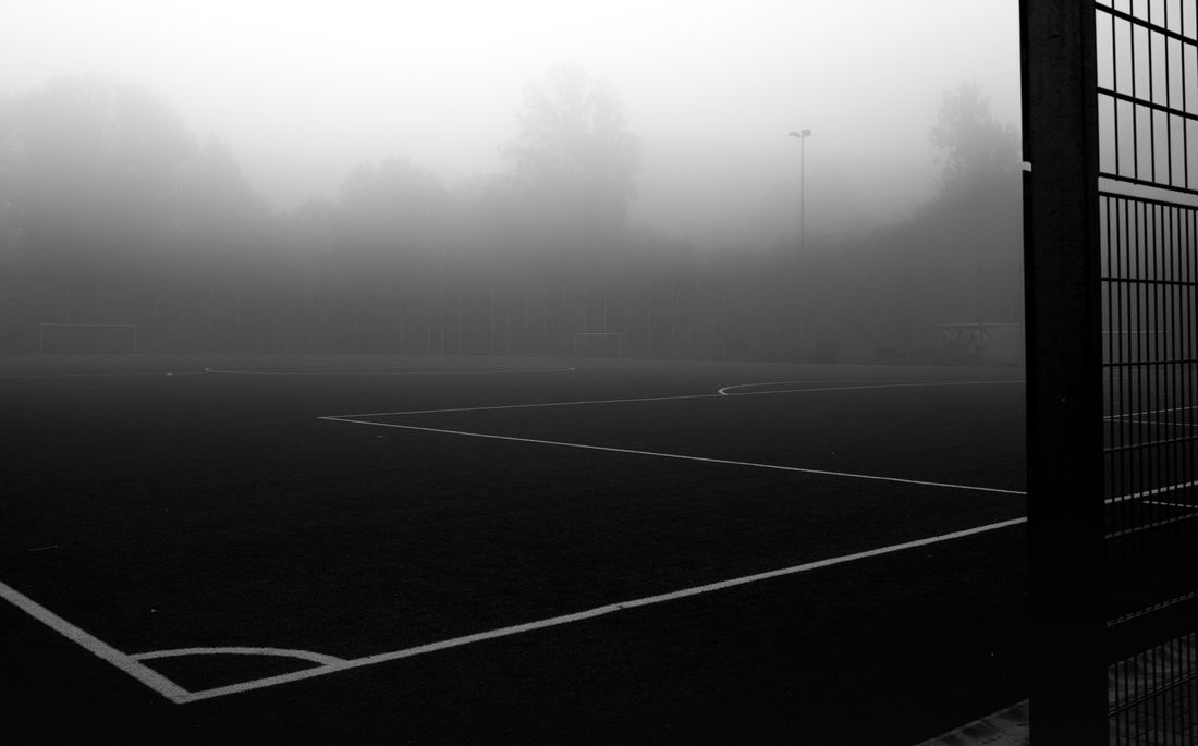 An empty football pitch in heavy fog, white lines on dark turf behind a fence