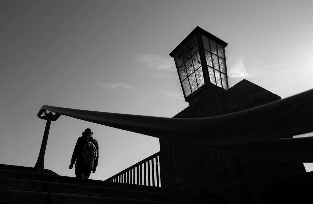 A walker in a hat climbs stairs below a curved handrail and a large lantern, seen against the sky
