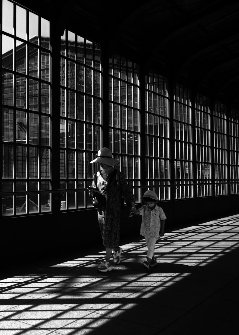 A woman in a sun hat walks hand in hand with a small child through a hall of tall windows and grid shadows