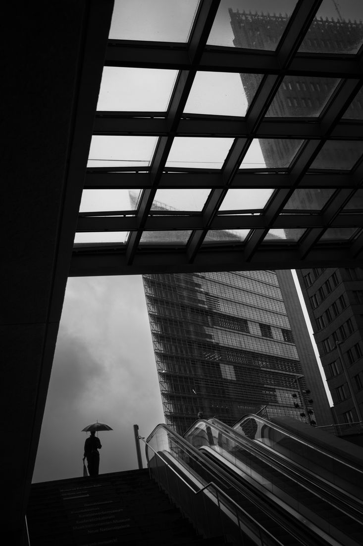 A figure with an umbrella stands at the top of stairs under a glass canopy, beside tall office towers