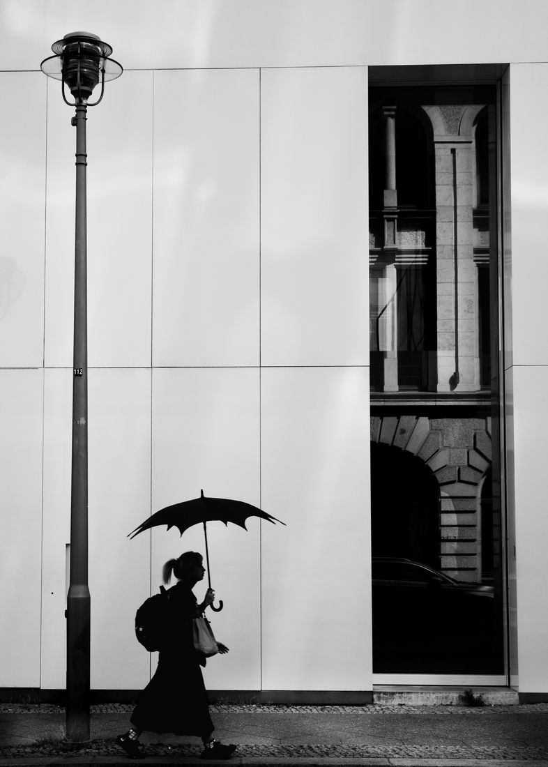 A woman with a scalloped umbrella walks past a white facade, beside a street lamp and a window reflecting an old stone building